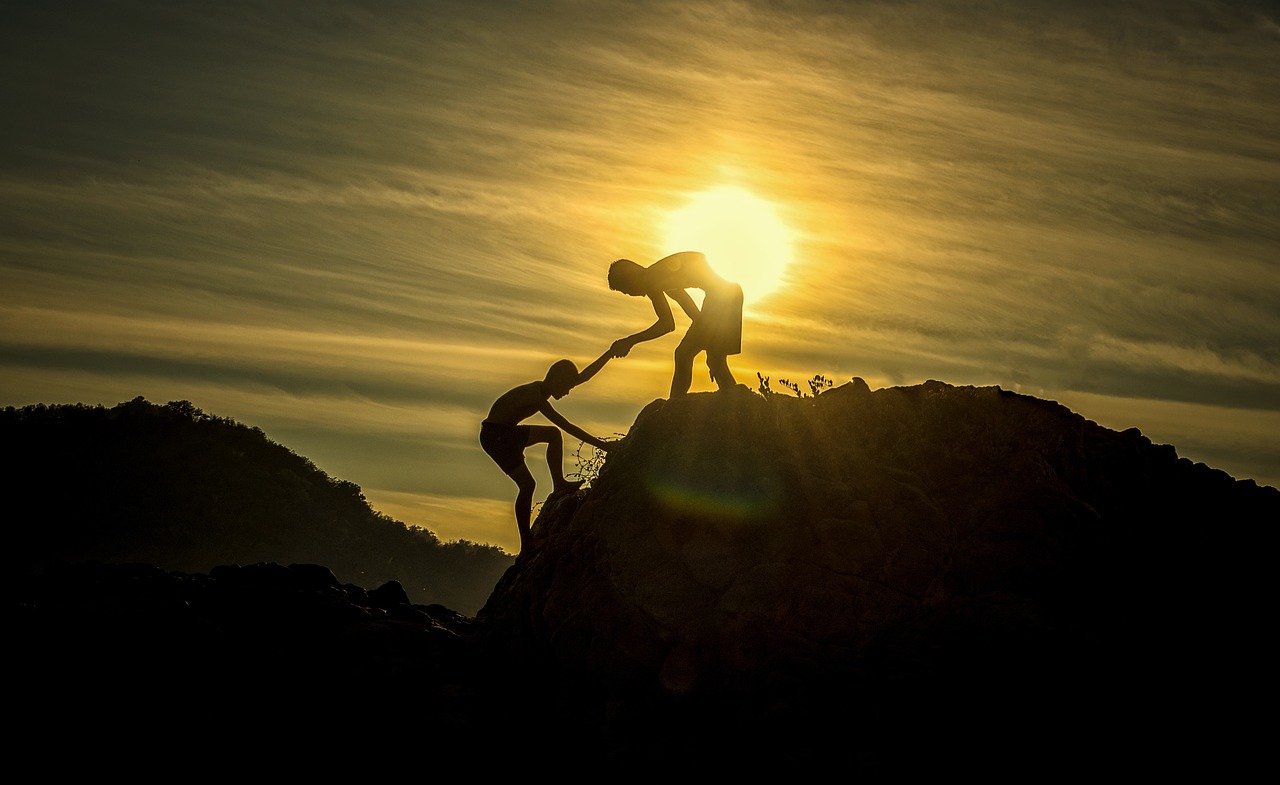 One person reaching down to help another climb a rocky summit at sunset